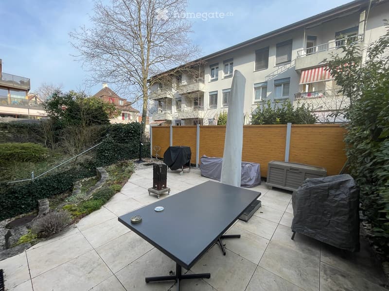 Garden with patio furniture, including a black table and covered chairs, surrounded by greenery and a wooden fence.