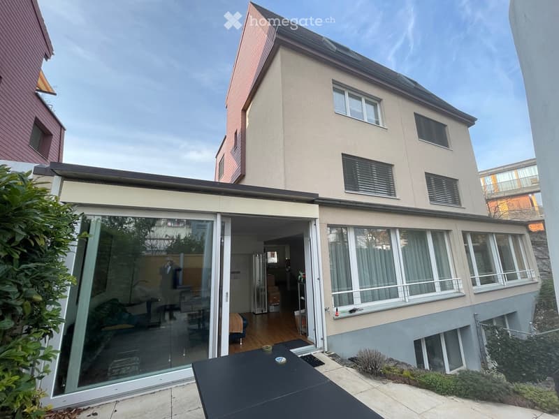 Modern two-story house with large windows, sliding glass doors leading to a patio, and a small garden area.