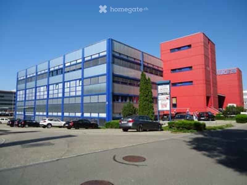 Two-story office building with red and blue exterior, multiple windows, and a parking lot in front.