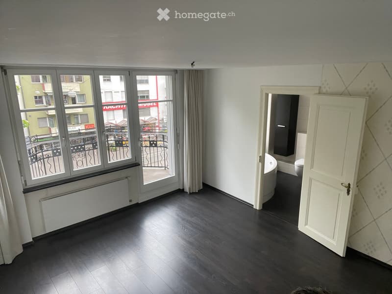 Empty room with dark wooden flooring, large windows with a view to a balcony, and an open door leading to a bathroom area.