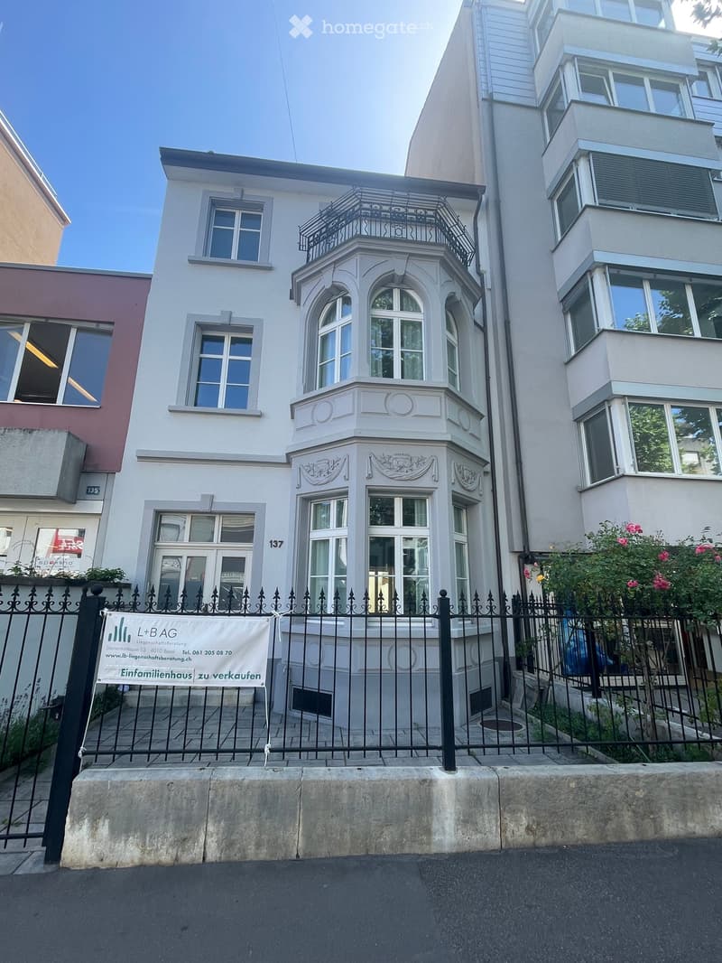 A three-story residential building with white facade, multiple windows, ornate architectural details, and a decorative balcony on the second floor.