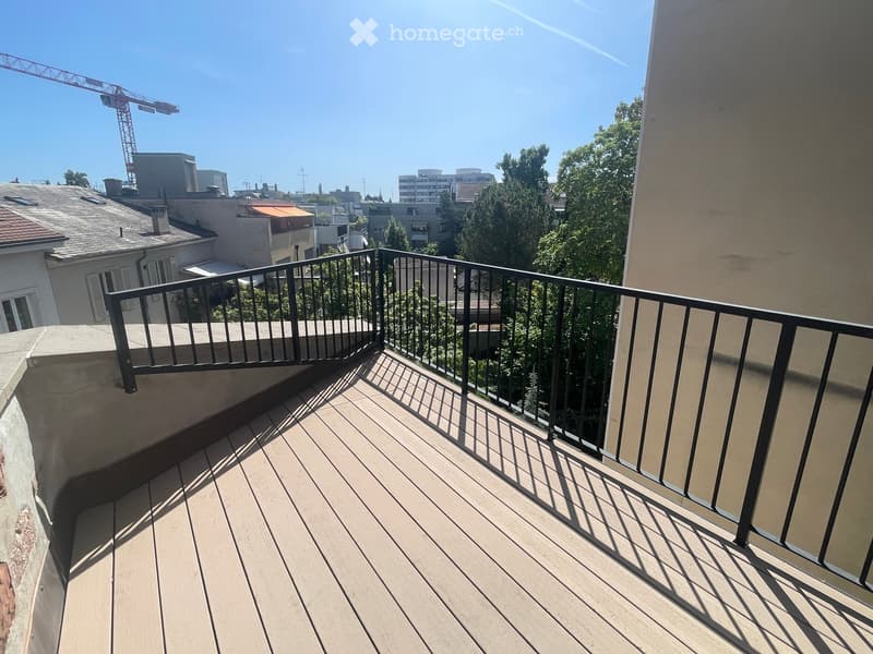 Balcony with wooden decking, black metal railing, overlooking a cityscape with buildings and trees.