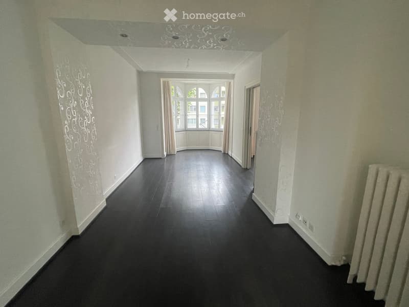 Empty hallway with dark flooring, light-colored walls, and a decorative ceiling design. A window at the end allows natural light to enter.