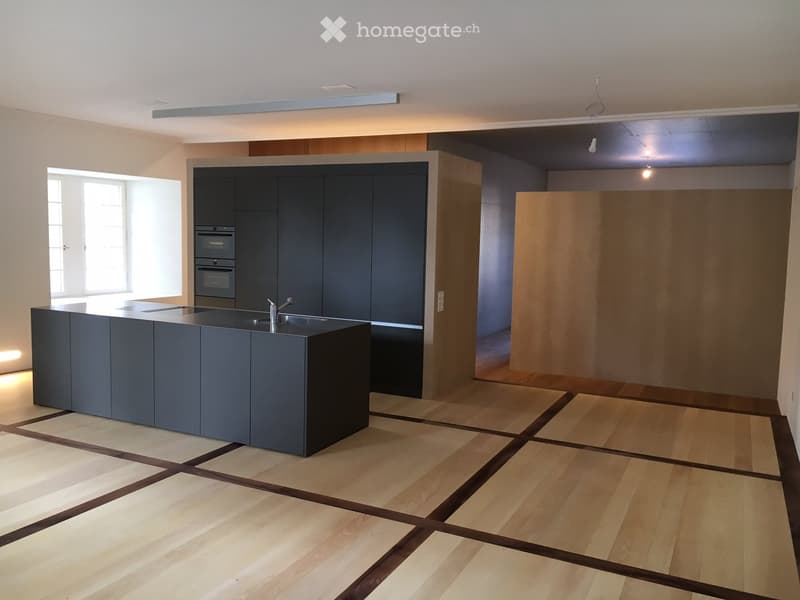 Modern kitchen with dark grey cabinets, a central island with a sink and faucet, and built-in ovens. The floor is made of light wooden planks arranged in a herringbone pattern.