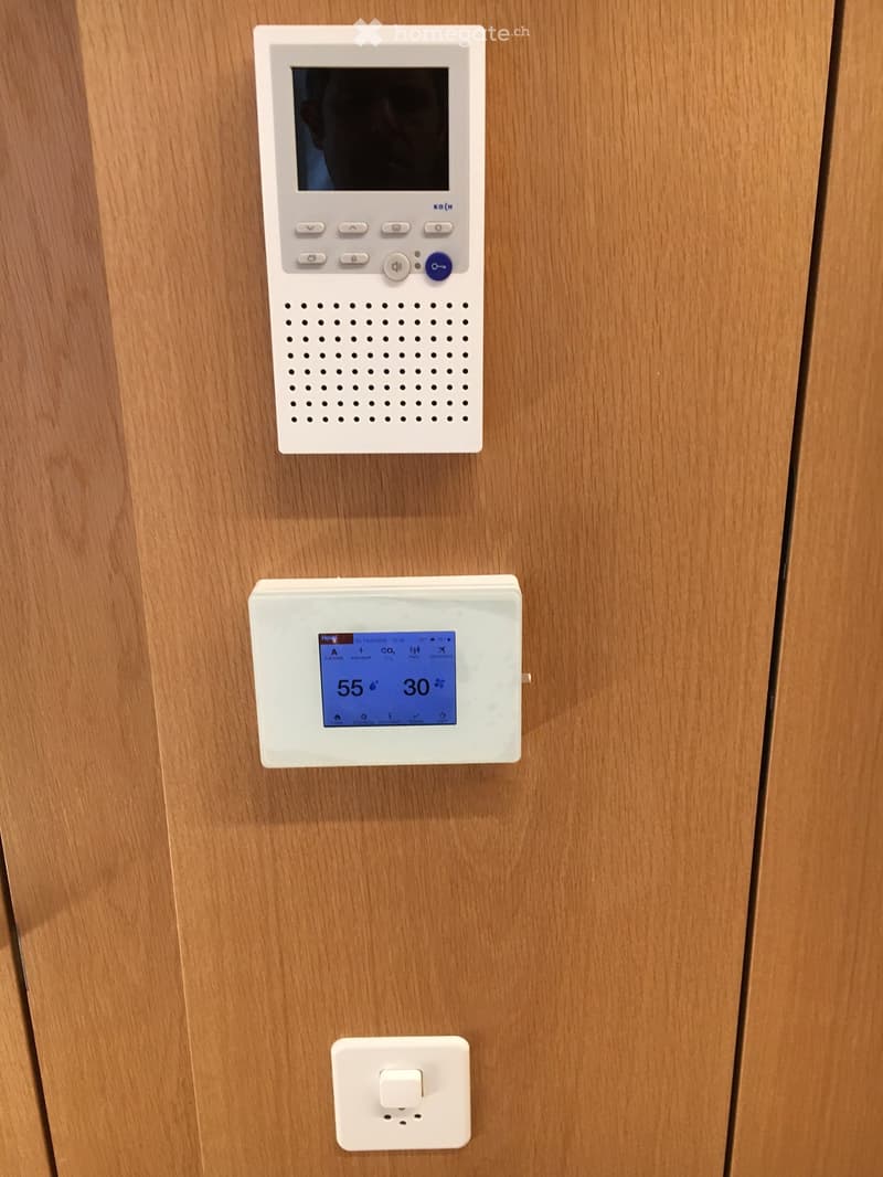 Wooden hallway with two white electronic devices mounted on the wall, one displaying temperature and humidity, and another with a screen and buttons.