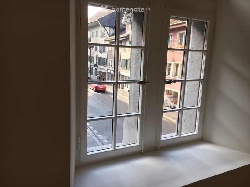 Empty room with large windows offering a view of a street with buildings and a red car passing by.