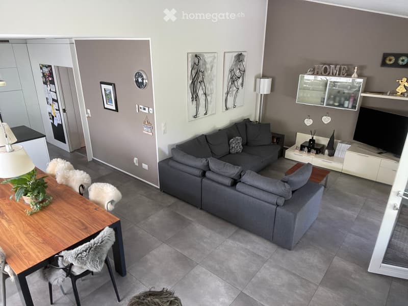 Modern living room with grey sectional sofa, wooden dining table, chairs, floor lamp, TV, shelves, and decorative items.