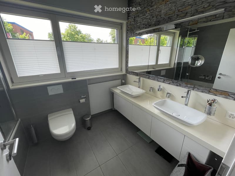 Modern bathroom with two sinks, a toilet, a large mirror, and a window with blinds. The walls are tiled in gray and stone, and the floor is tiled in dark gray.