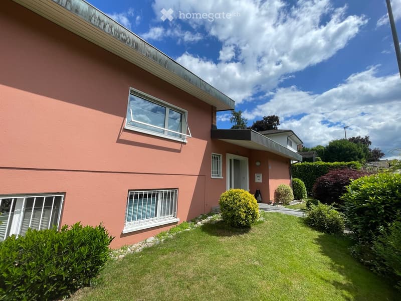 Two-story pink house with white-framed windows, a small balcony, and a well-maintained garden with green grass and trimmed bushes.