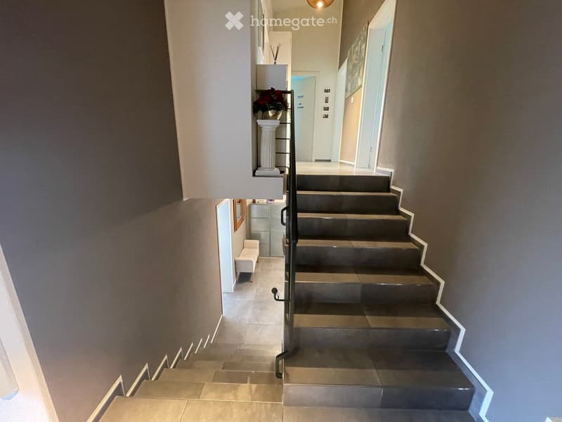 Spacious hallway with modern staircase, grey walls, and decorative items including a small bench and a vase with flowers on a shelf.