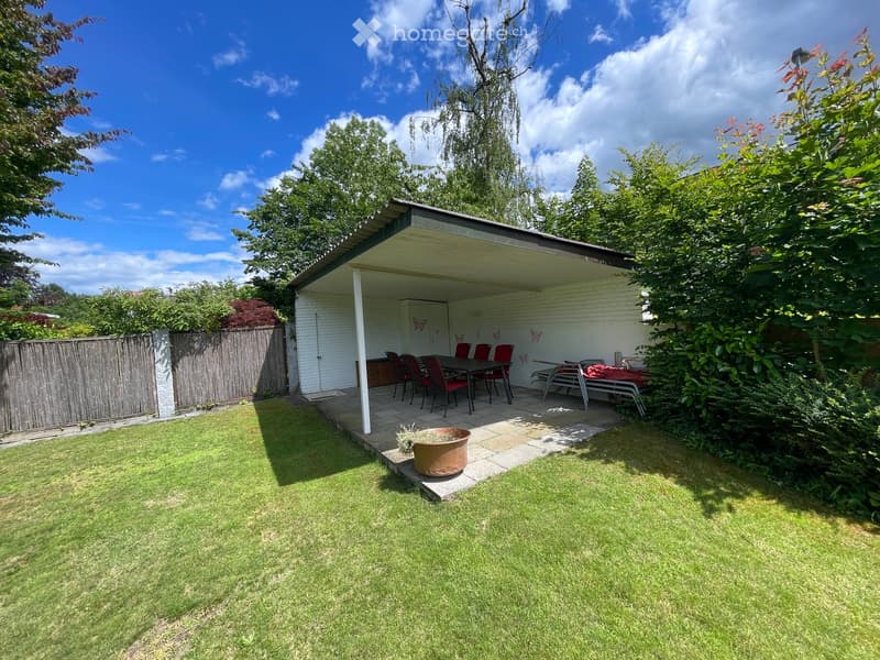 A well-maintained garden with a grassy lawn, a covered patio area with a table and chairs, and a large flower pot near the patio.