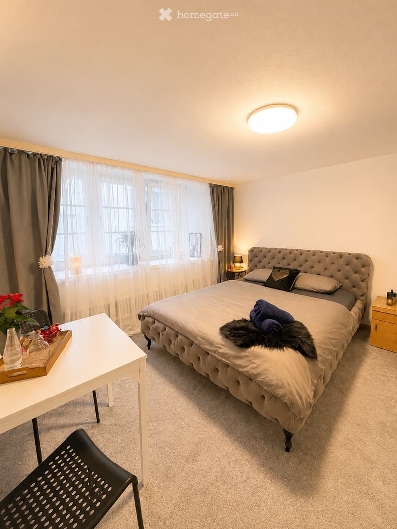 Well-lit bedroom with a large, tufted headboard bed, nightstand, side table, and decorative items including a flower vase and a tray with glassware.