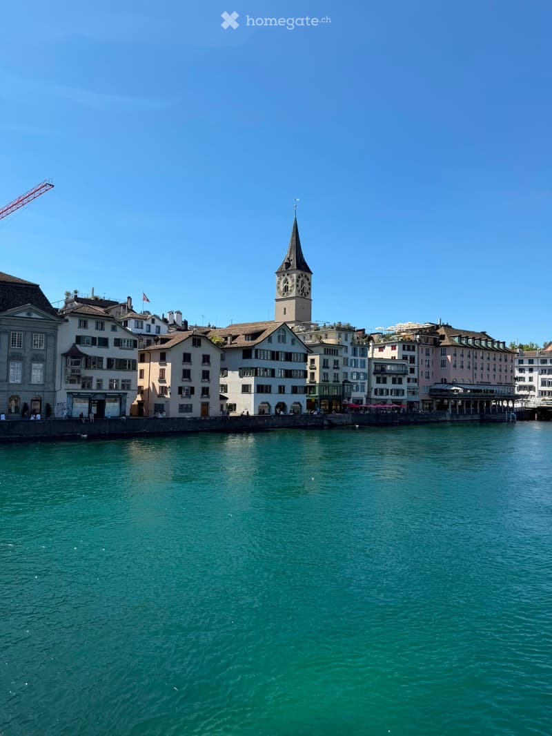A view of a cityscape with a prominent clock tower overlooking a calm river, surrounded by multi-story buildings with various architectural styles.