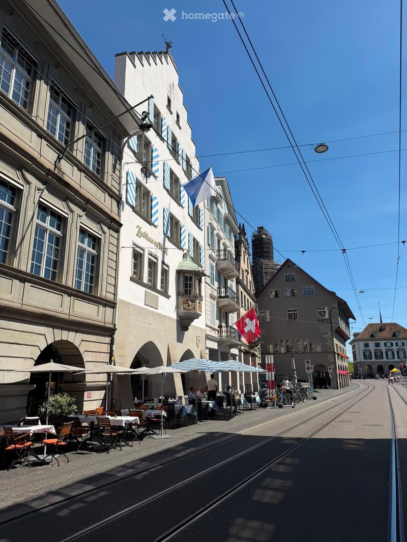 Outdoor seating area with tables, chairs, and umbrellas in front of a historic building with Swiss flags and a tram track running alongside.