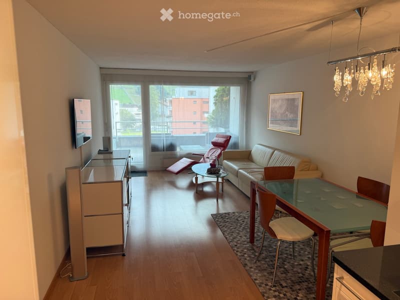 Modern living room with a sofa, coffee table, dining table, chairs, and a TV screen. Large windows provide natural light, and the space features wooden flooring and contemporary decor.