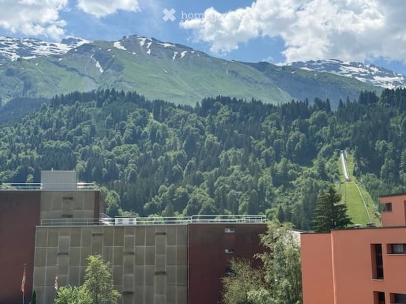 Modern building with large glass windows and a flat roof, situated in a lush green valley with snow-capped mountains in the background.