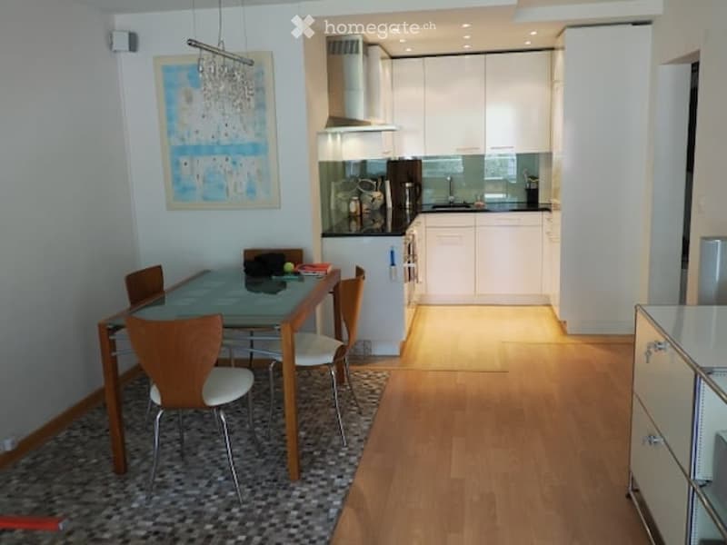 Modern open-plan kitchen and dining area with light-colored cabinets, a glass dining table with four chairs, and a sleek stove. The space features wooden flooring and a contemporary light fixture.