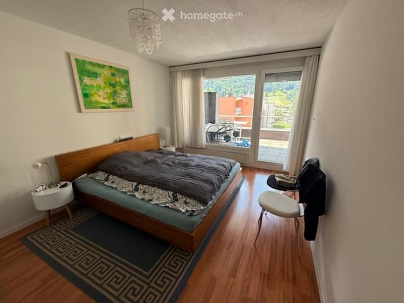 Cozy bedroom with wooden flooring, a large bed with dark bedding, a nightstand, a chair, and a painting on the wall. Large windows with curtains lead to a balcony.
