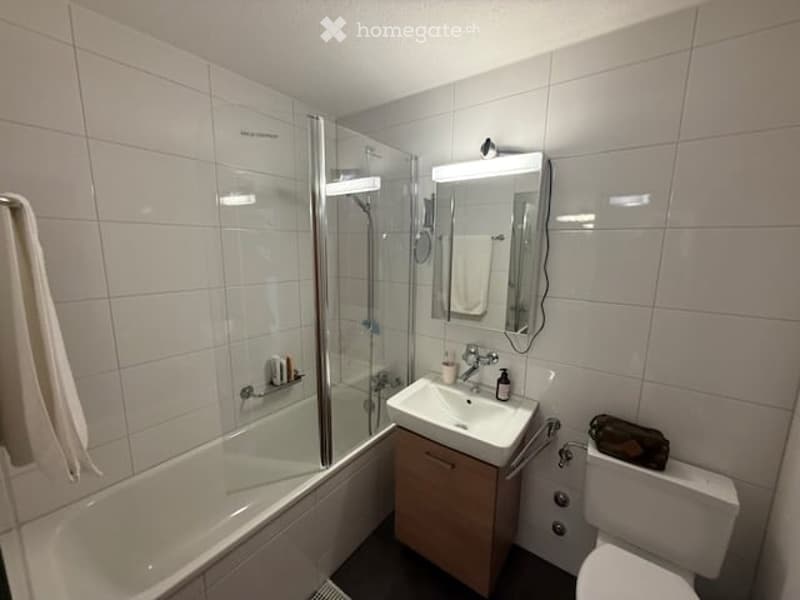 Modern bathroom with white tiles, a bathtub, a sink with a mirror, and a toilet. A towel hangs on the side of the bathtub, and a small bag is placed on the toilet seat.
