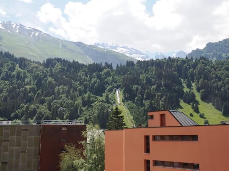 View from a balcony or terrace showing a modern building complex with orange and brown facades, surrounded by lush green forest and snow-capped mountains in the background.