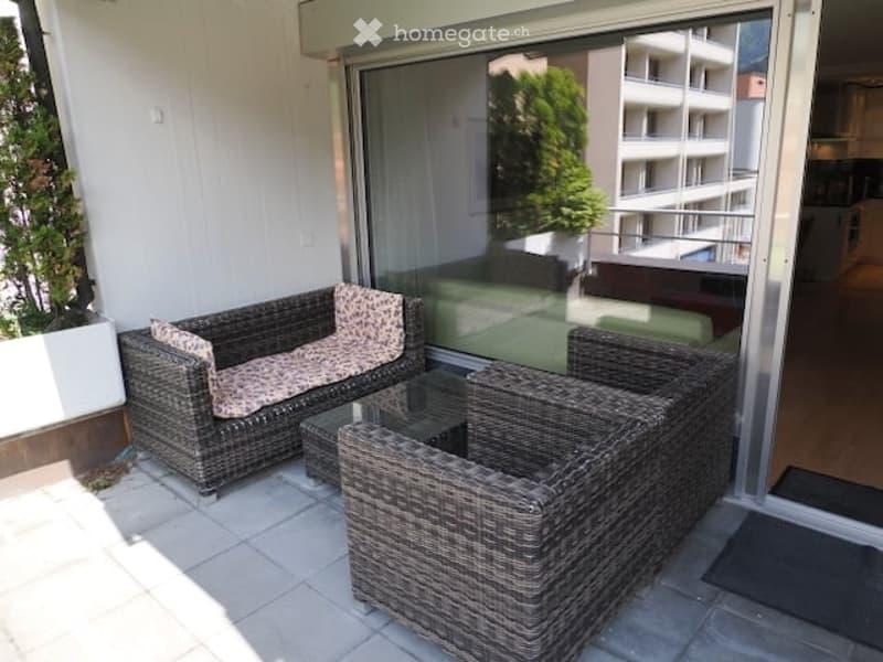 Balcony with wicker furniture including a sofa, coffee table, and chairs, overlooking a cityscape with modern buildings and greenery.
