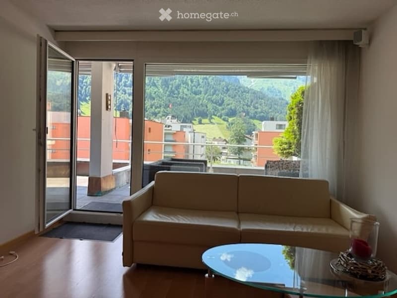 Bright living room with large windows offering a scenic mountain view, featuring a cream-colored sofa, a glass coffee table, and a chair near the balcony entrance.