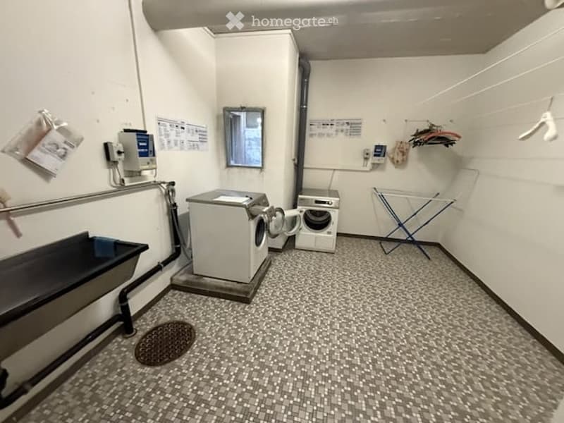 Laundry room with a washing machine, sink, mirror, and a drying rack. The room has tiled flooring and white walls.