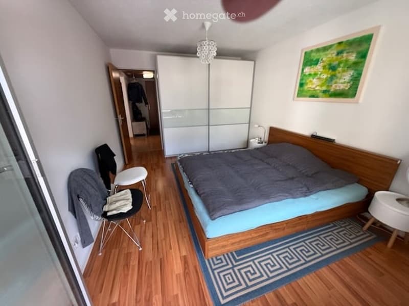 Cozy bedroom with wooden flooring, a double bed with grey bedding, a white wardrobe, a chair, a small stool, and a decorative picture frame on the wall.