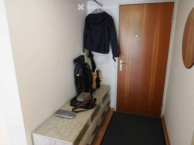 A small hallway with a wooden bench holding a backpack, handbag, jacket, and umbrella, next to a closed wooden door.