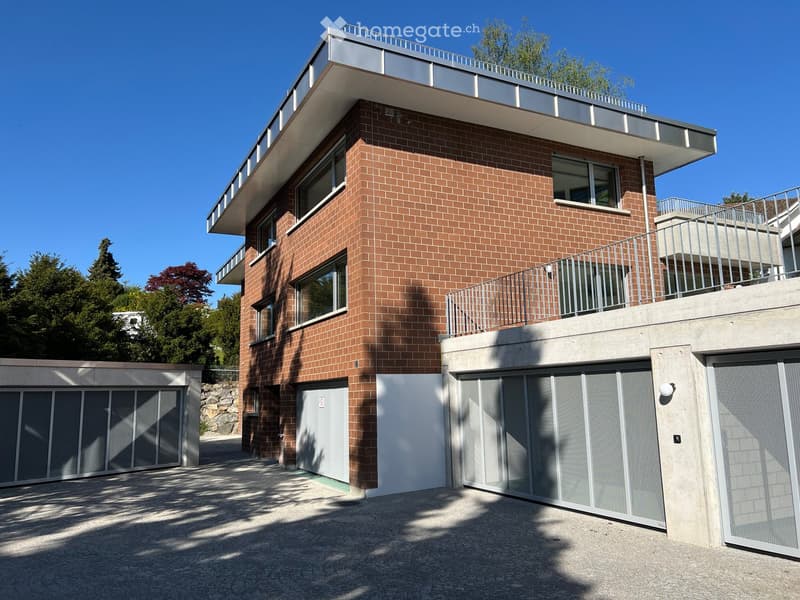 Modern two-story building with red brick facade, large windows, and balconies. Features a garage with multiple car doors and a clean, contemporary design.