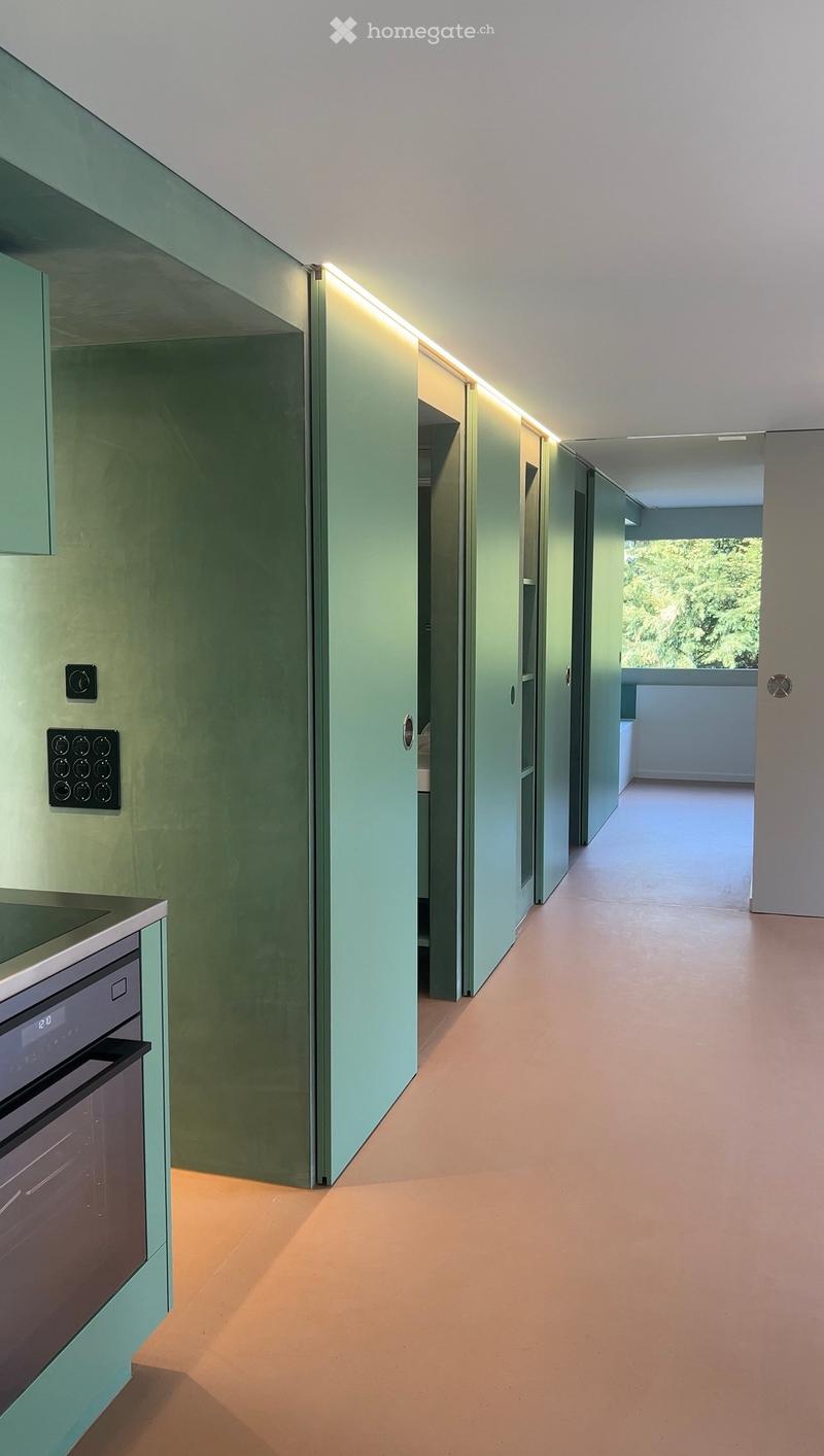 Modern hallway with light-colored walls, sleek cabinetry, and recessed lighting. Features a built-in oven and a view of a balcony area with greenery.