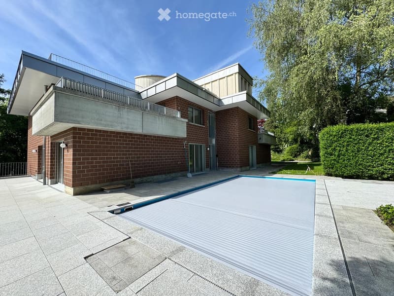Modern two-story house with brick and concrete exterior, large balcony, swimming pool, and landscaped garden.