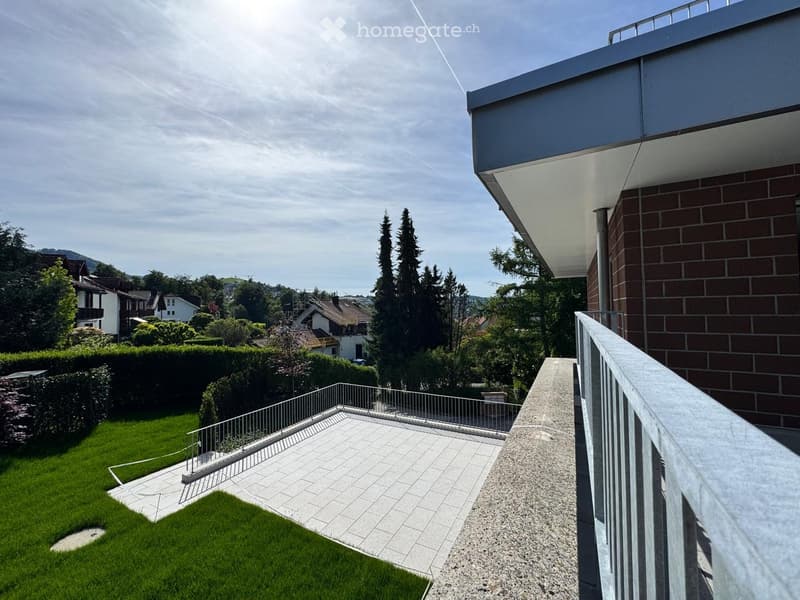 Balcony overlooking a well-maintained garden with a tiled patio area, surrounded by trees and neighboring houses.