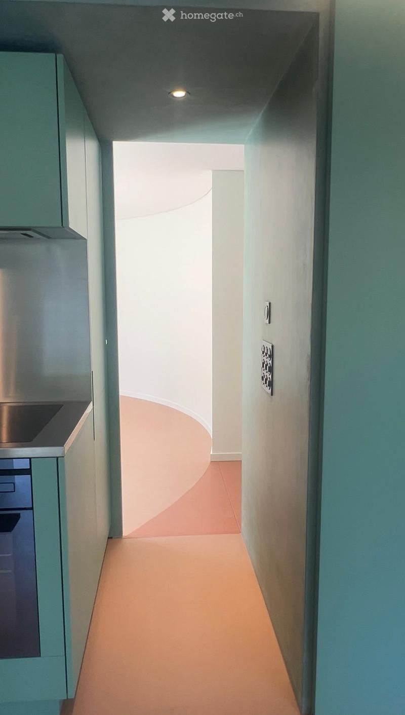Modern hallway with light-colored walls, a curved ceiling, and a warm-toned floor. A small section of a kitchen with cabinets and a countertop is visible on the left side.