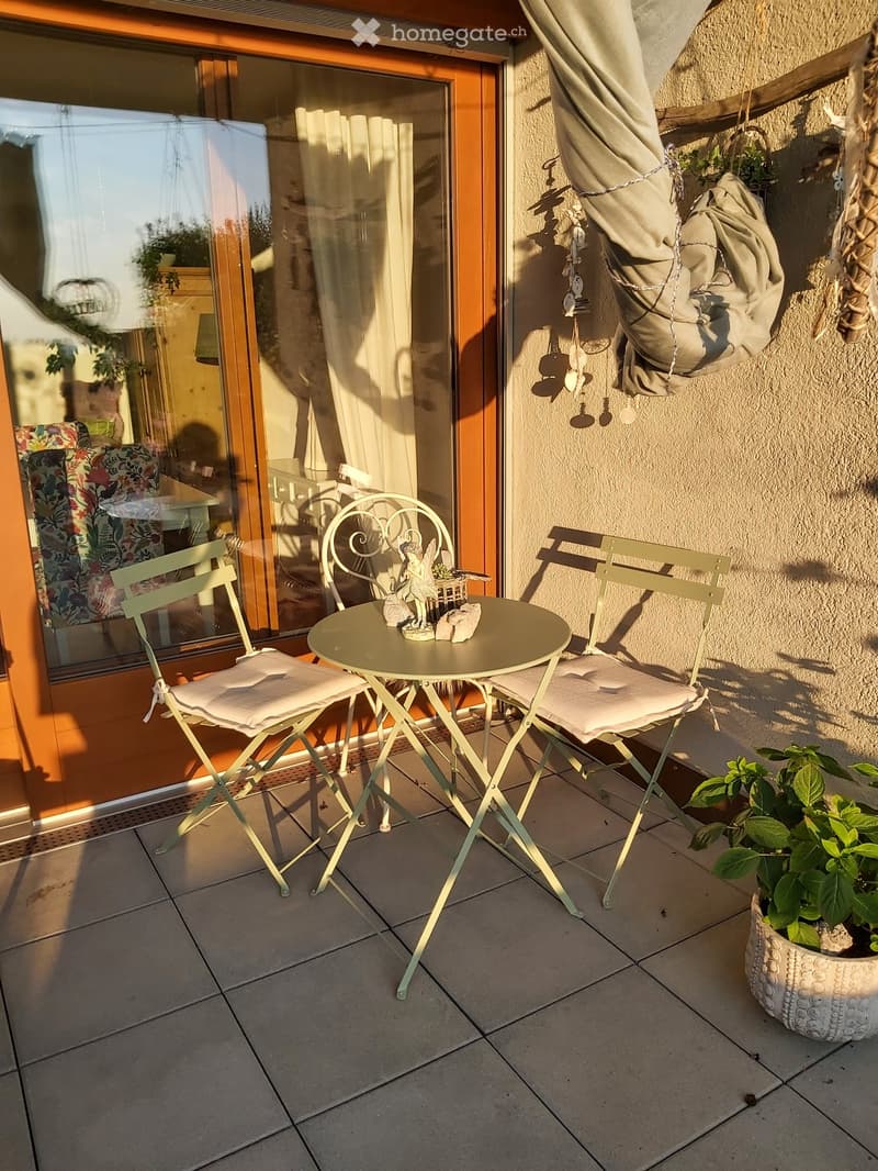 Balcony with a small round table, two chairs, a decorative statue, and a potted plant. The balcony has tiled flooring and is adjacent to a glass door leading inside.