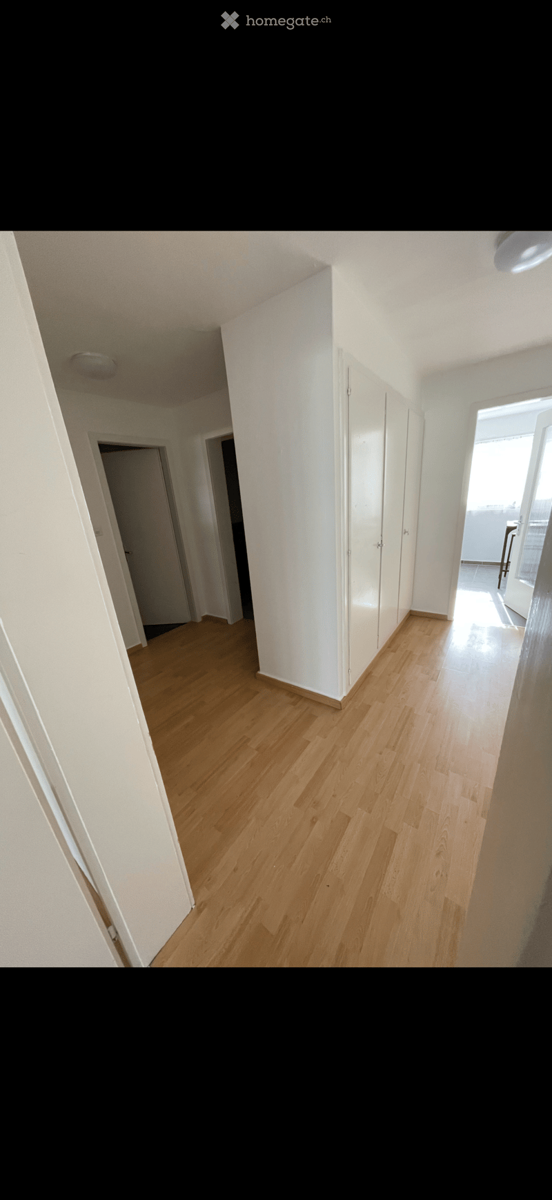 Bright, empty hallway with light wooden flooring and white walls, featuring several doors and a visible room at the end.
