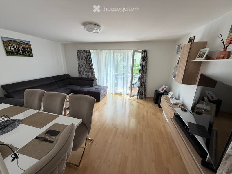 Modern living and dining area with a grey sectional sofa, white dining table with four chairs, TV stand, shelves, and a balcony access.