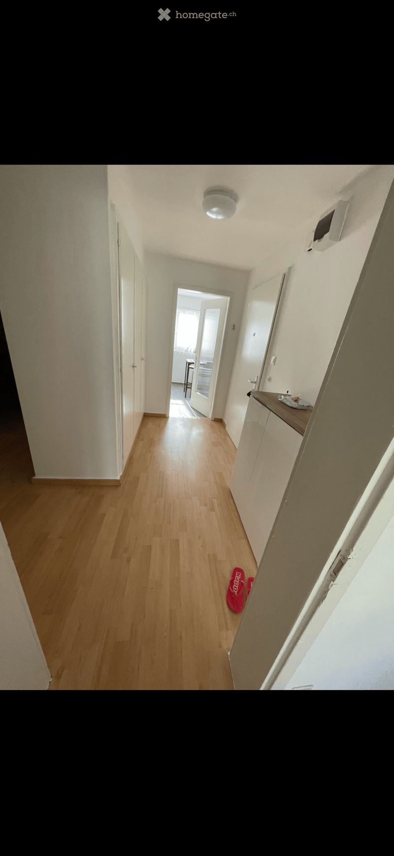 A narrow hallway with wooden flooring, white walls, and a ceiling light. A pair of red slippers is placed on the floor near a small table with a countertop and a few items on it. The hallway leads to an open doorway revealing a glimpse of another room wit