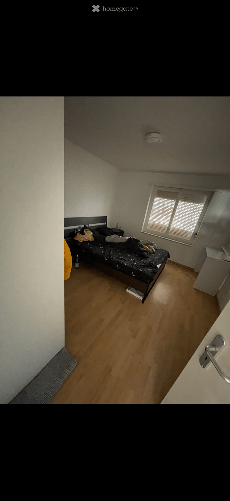 Bedroom with wooden floor, bed with dark bedding, nightstand, and a window with blinds.