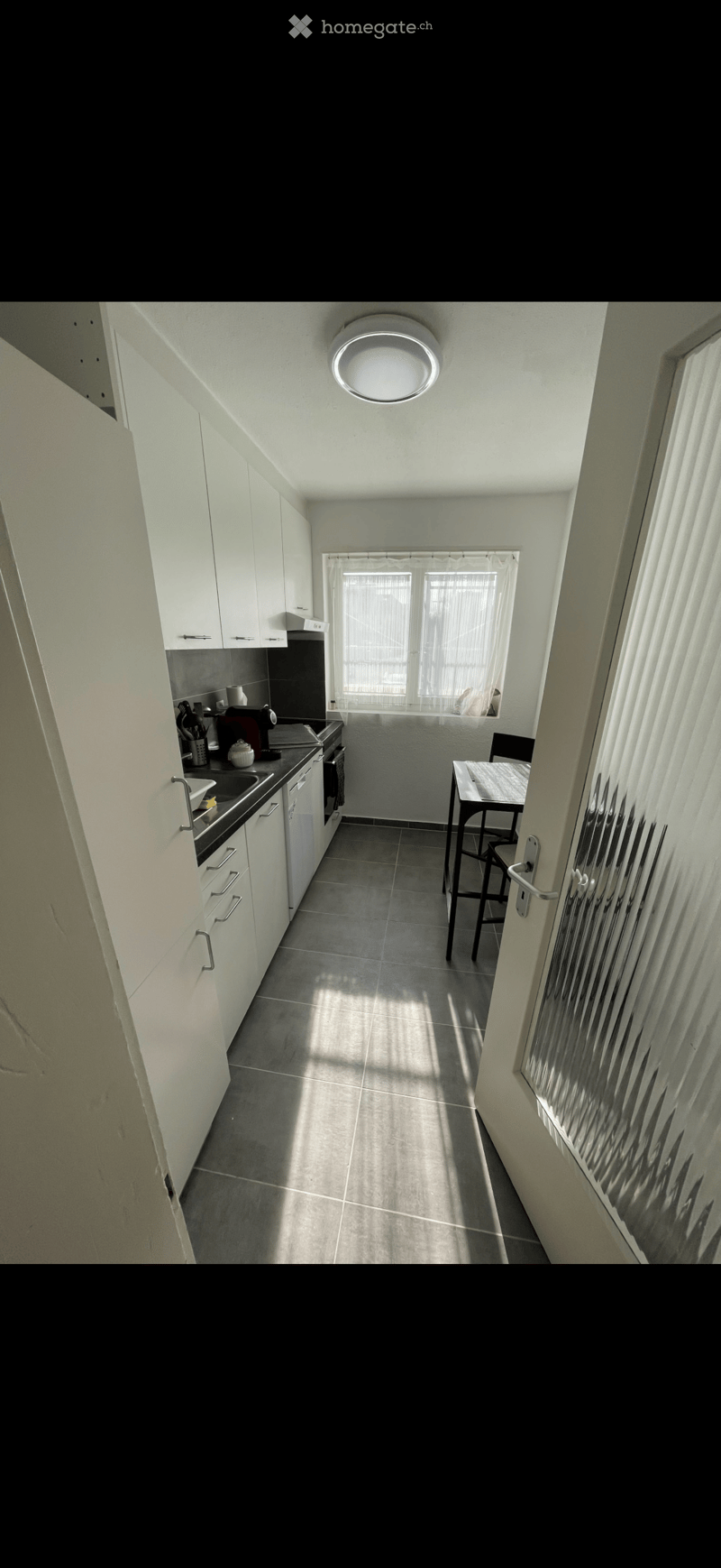 Modern kitchen with white cabinets, dark countertops, and a small dining area with a table and chairs. Natural light streams in from a window with blinds.
