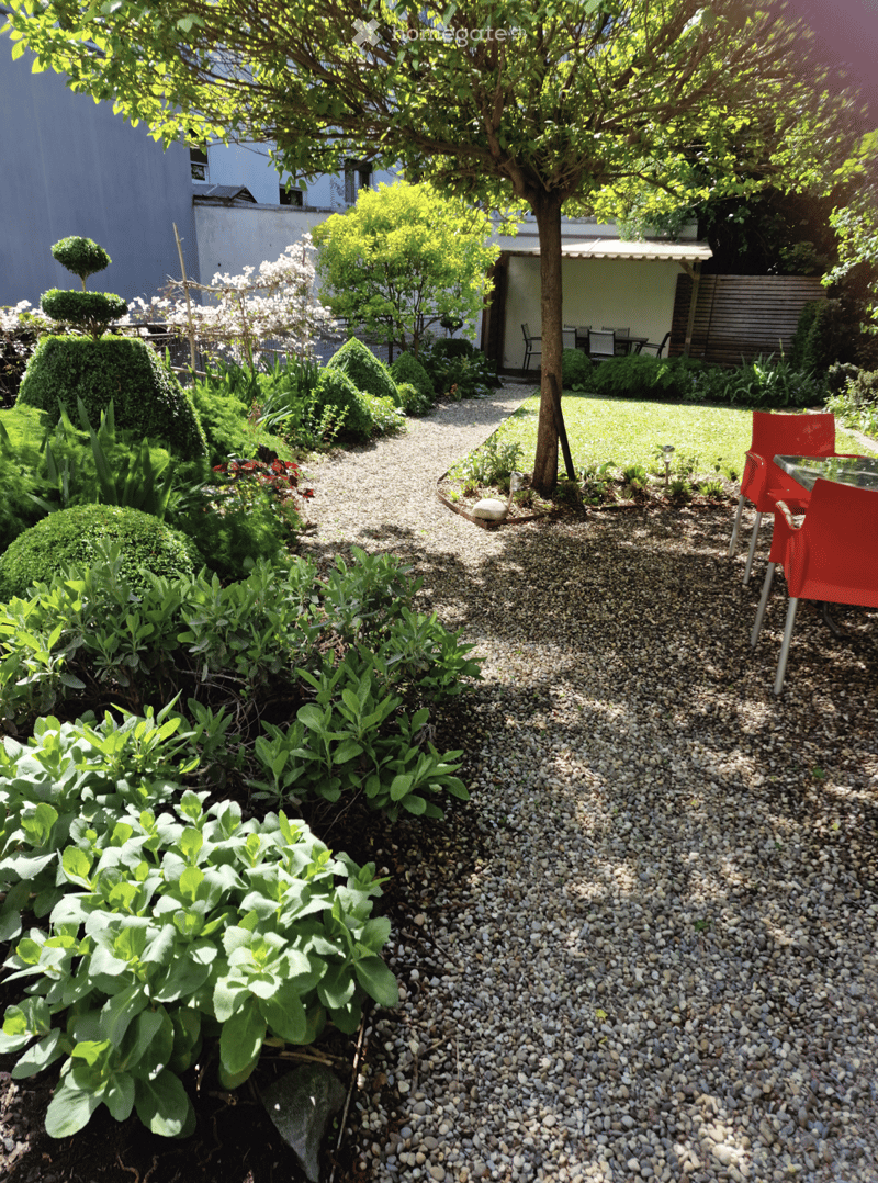 Well-maintained garden with gravel pathway, trimmed shrubs, flowering trees, and outdoor seating area with red chairs and a table.