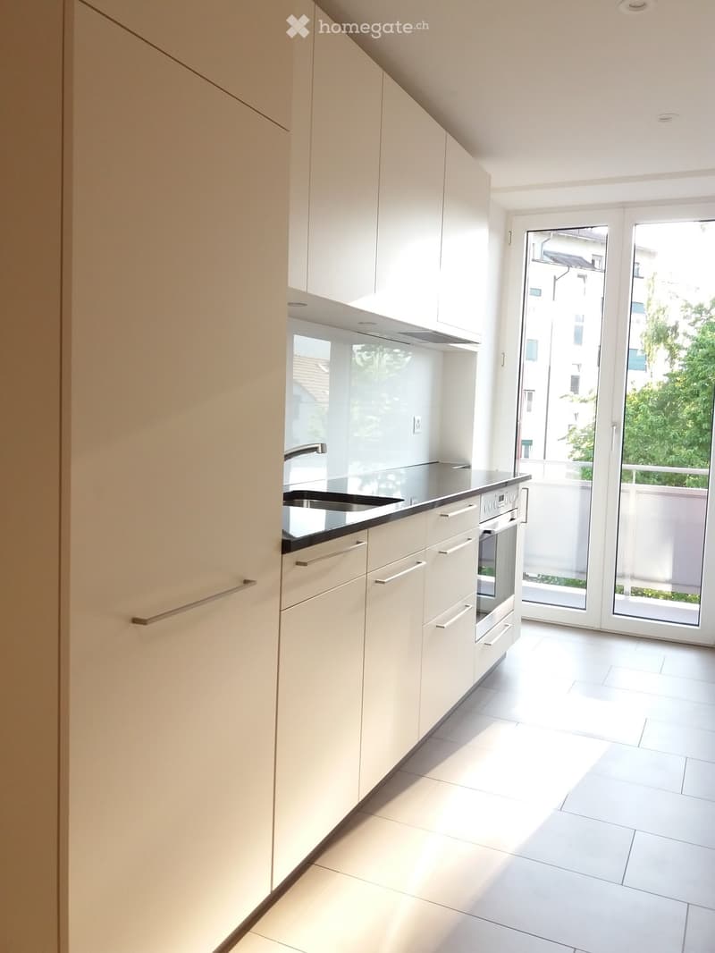 Modern kitchen with white cabinets, a black countertop, a sink, and an oven. The space is bright with large windows and a sliding glass door leading to a balcony.