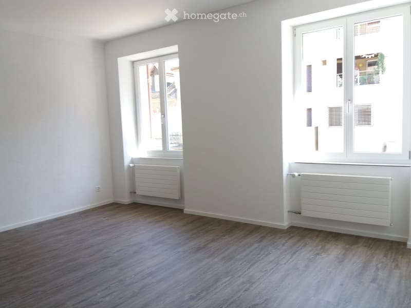 Empty room with light-colored walls, wooden flooring, and two radiators. Windows on two walls provide natural light.
