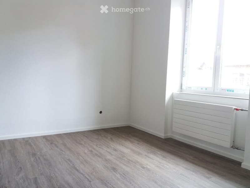 Empty room with white walls, wooden flooring, and a window with a radiator below it.