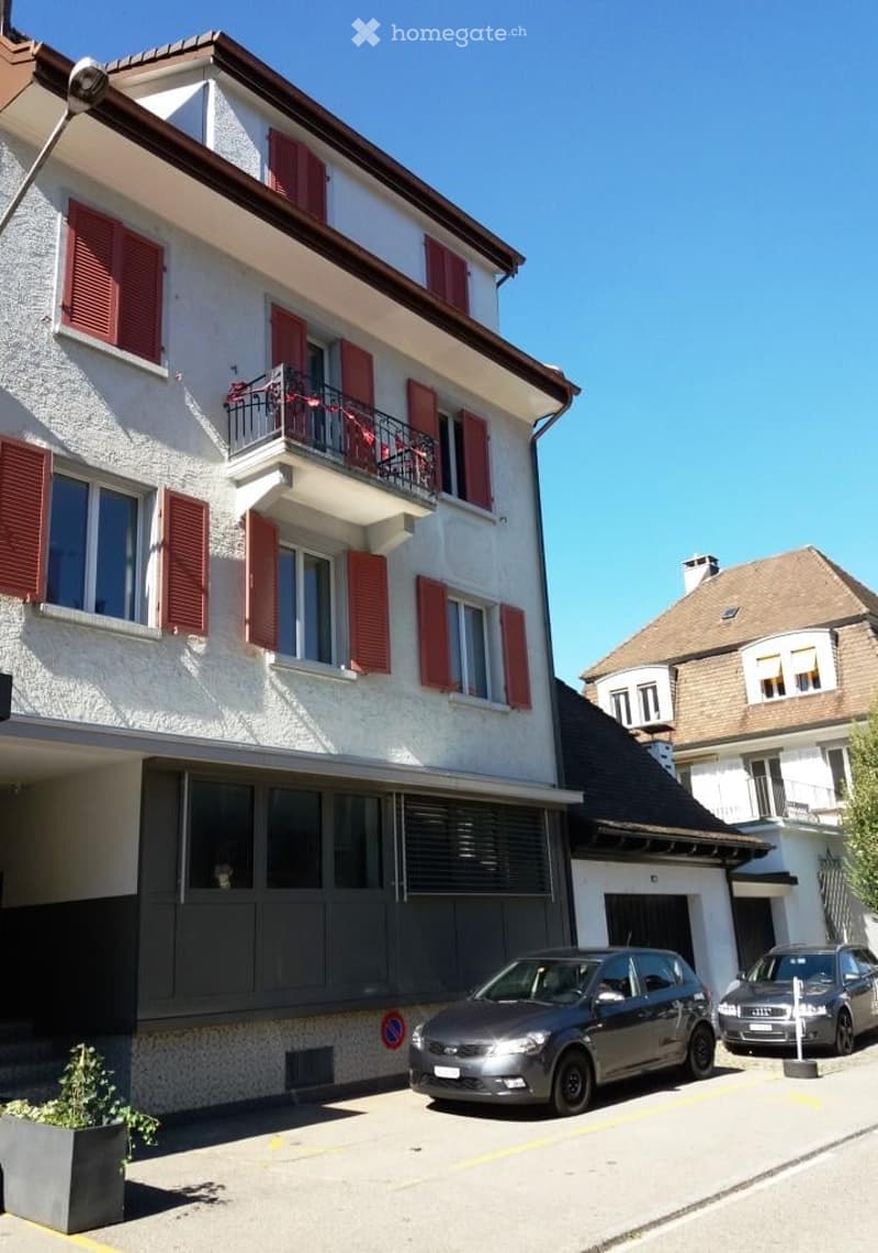 Multi-story residential building with white facade, red shutters, and a balcony on the second floor. A covered garage and parking area are visible in front.