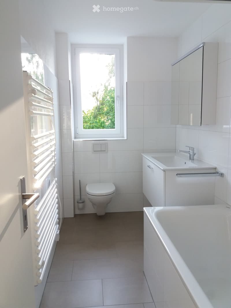 Modern bathroom with white tiles, a toilet, a bathtub, a sink with a faucet, a mirror, and a radiator.