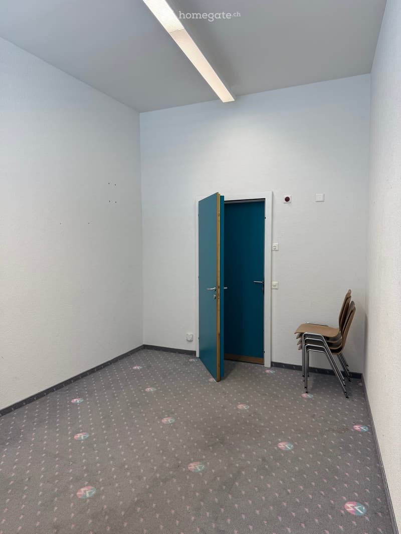 Empty room with light blue walls, grey carpet with colorful patterns, and a single stack of chairs in the corner.