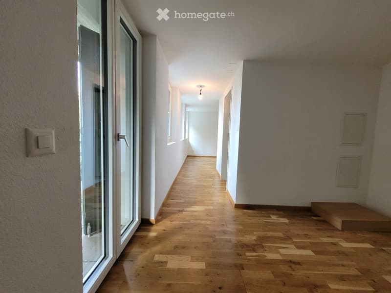 Empty hallway with wooden flooring, white walls, and a large glass door on the left side. The hallway is well-lit with recessed ceiling lights and has a small step leading to another area at the end.