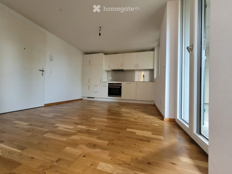 Empty room with light wooden flooring, white walls, and a small kitchen area with cabinets and appliances.