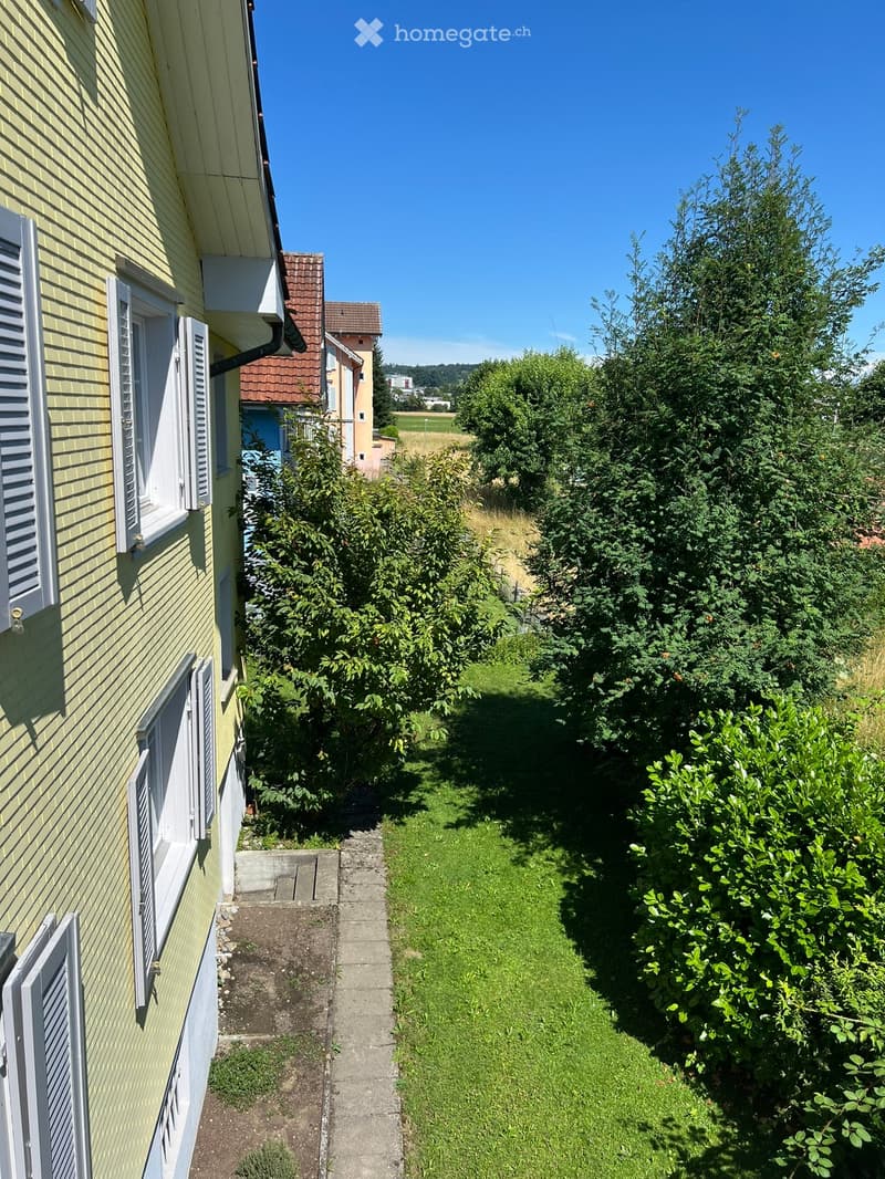 Two-story house with yellow exterior, white windows with shutters, adjacent building, garden, greenery, pathway